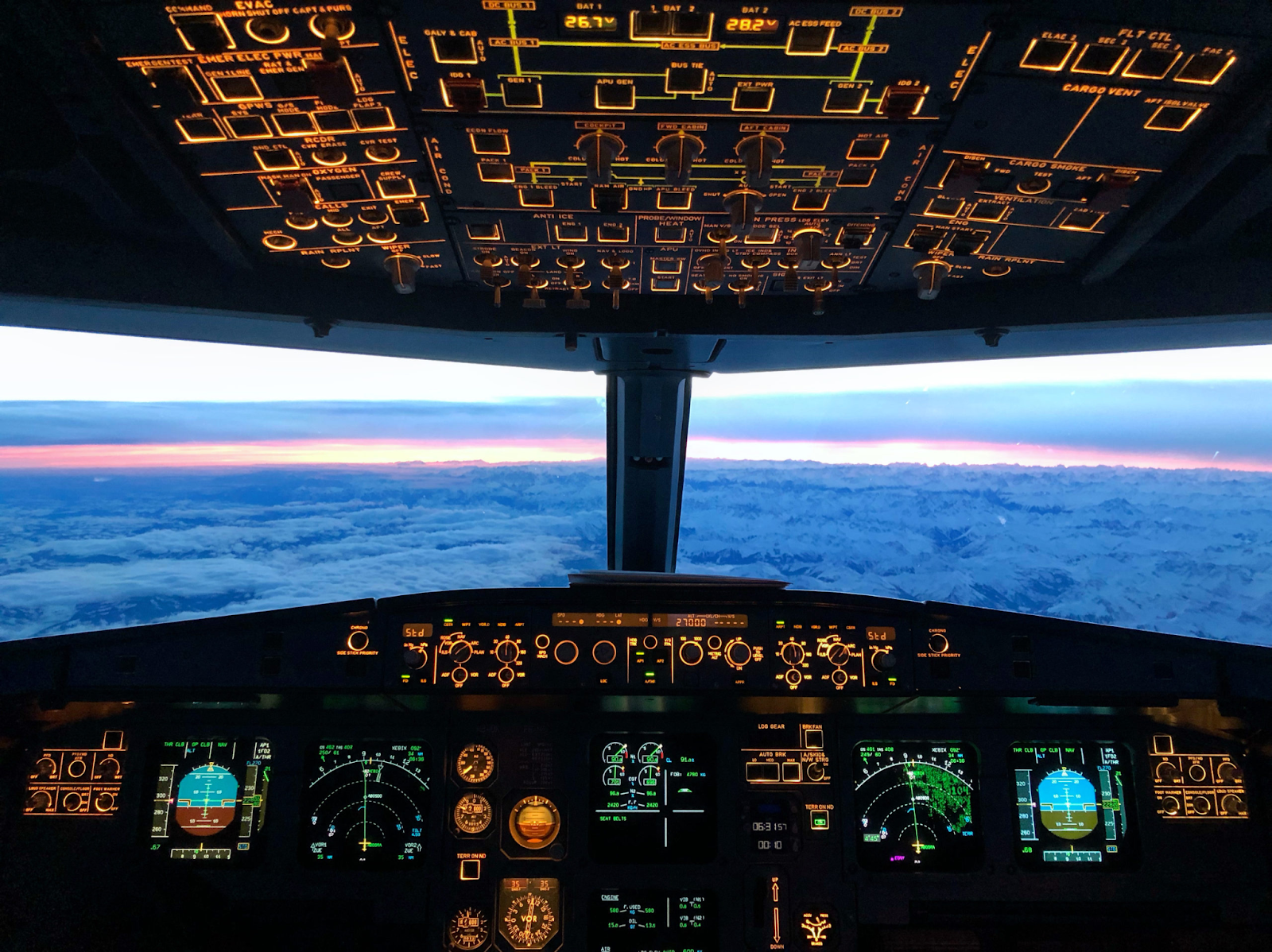 Cockpit view with illuminated controls and displays, overlooking snowy mountains and a colorful sunset sky